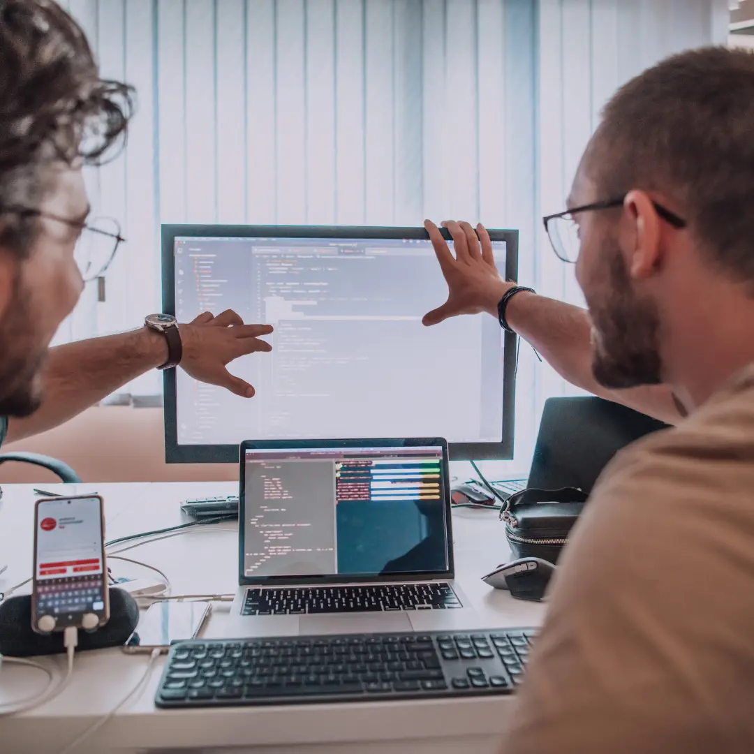 Developers collaborating on code displayed on a desktop monitor and a laptop, showcasing seamless integration with major platforms like Shopify and WooCommerce, emphasizing teamwork and technical expertise in secure digital content delivery.